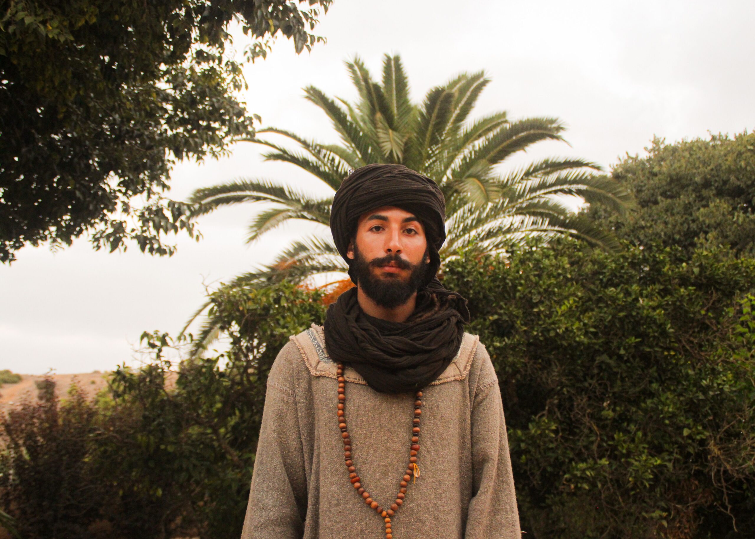 Portrait of a Moroccan man in traditional attire standing outdoors with lush greenery.